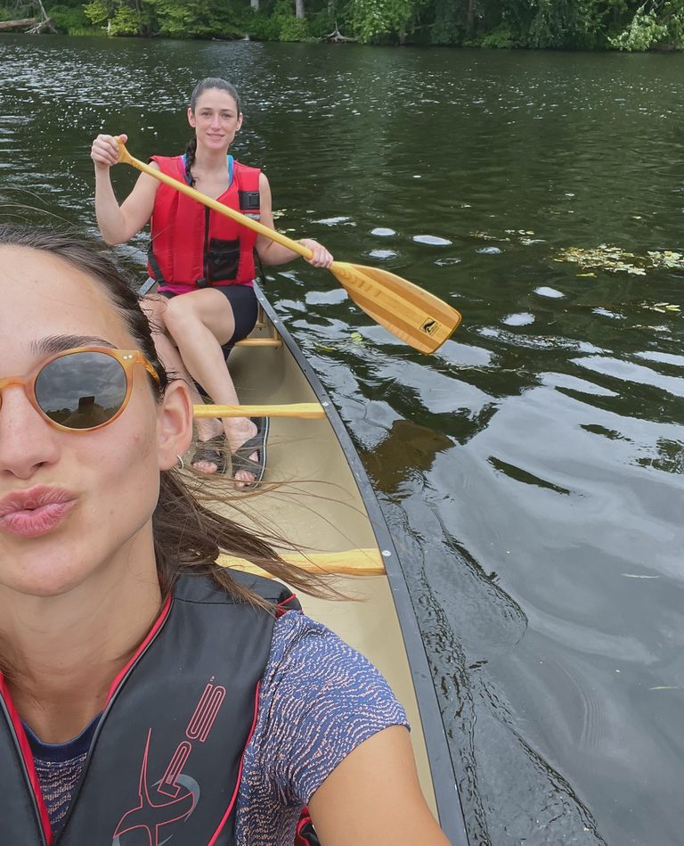 Two women in life jackets in a canoe on a dark green lake; the one in the stern holds a wooden paddle mid-stroke.