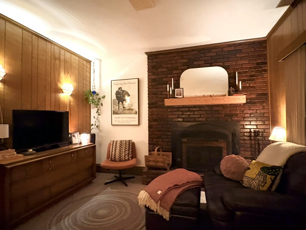A panelled family room at night: a vintage wooden credenza below the television, two lit wall sconces, a framed Brandywine River Museum poster, a salmon swivel chair and a brick fireplace with a mirror above the mantel.