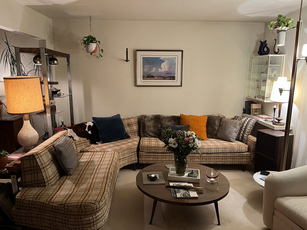 A living room with a plaid mid-century sectional, a large ceramic table lamp, a round wooden coffee table with flowers, and a framed landscape print above the sofa.