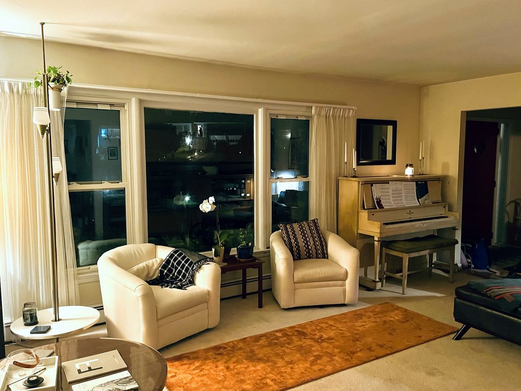 Two cream barrel chairs in front of a wide window at night, a rust-coloured rug, and an upright piano with candles and sheet music.