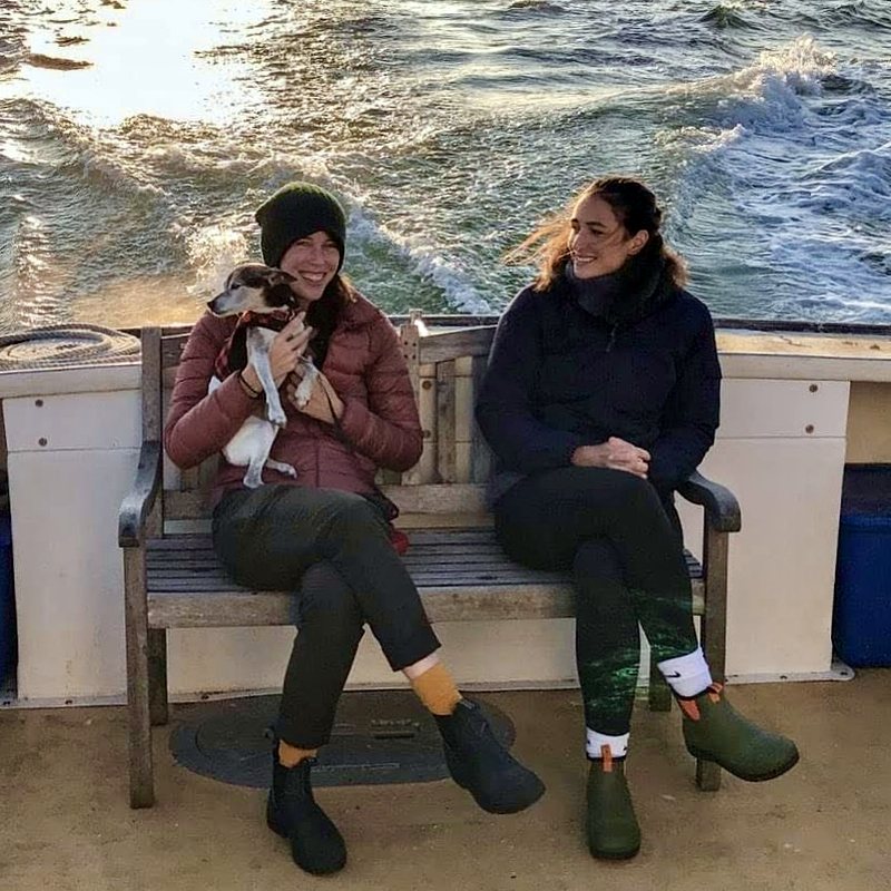 Elizabeth and a friend sitting on a bench at the stern of a boat in cold weather, waves behind them, a small dog on Elizabeth's lap.